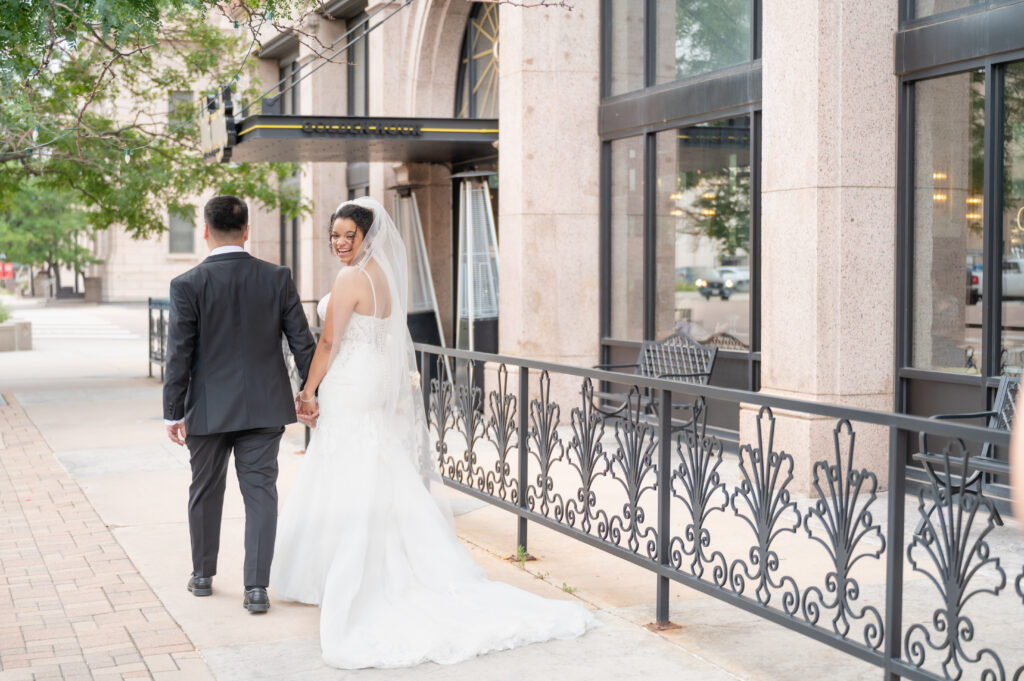 Bride looks over her shoulder and smiles as she strolls hand in hand with her new husband through the heart of downtown Colorado Springs