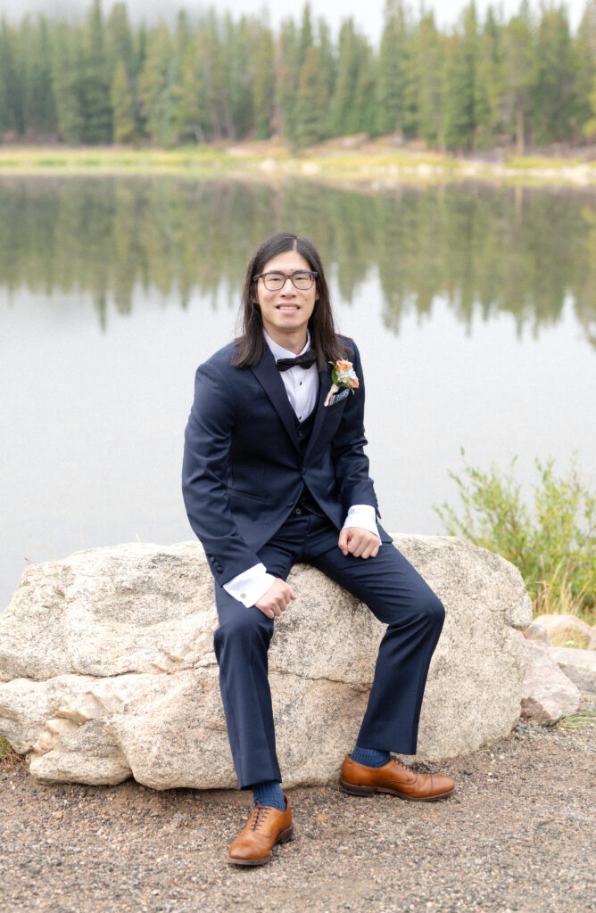 Groom in blue suit seated on a rock with Echo Lake in the background on his elopement morning