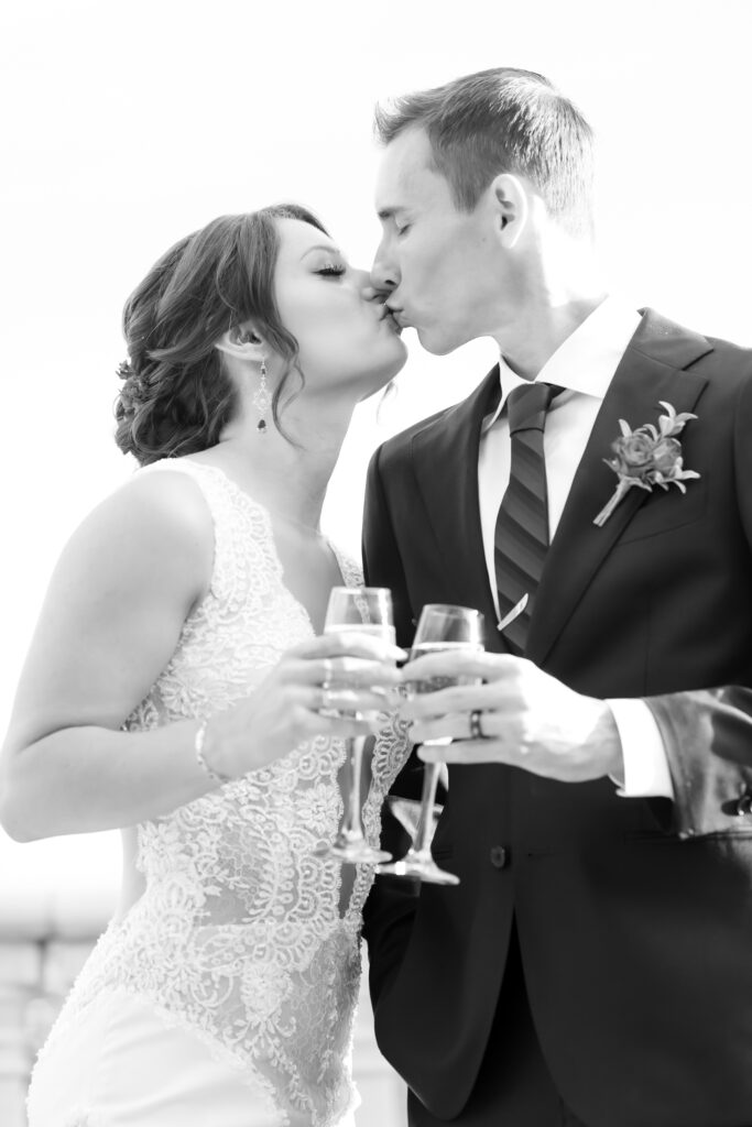 Black and white image of the bride and groom kissing and toasting their glasses of champagne