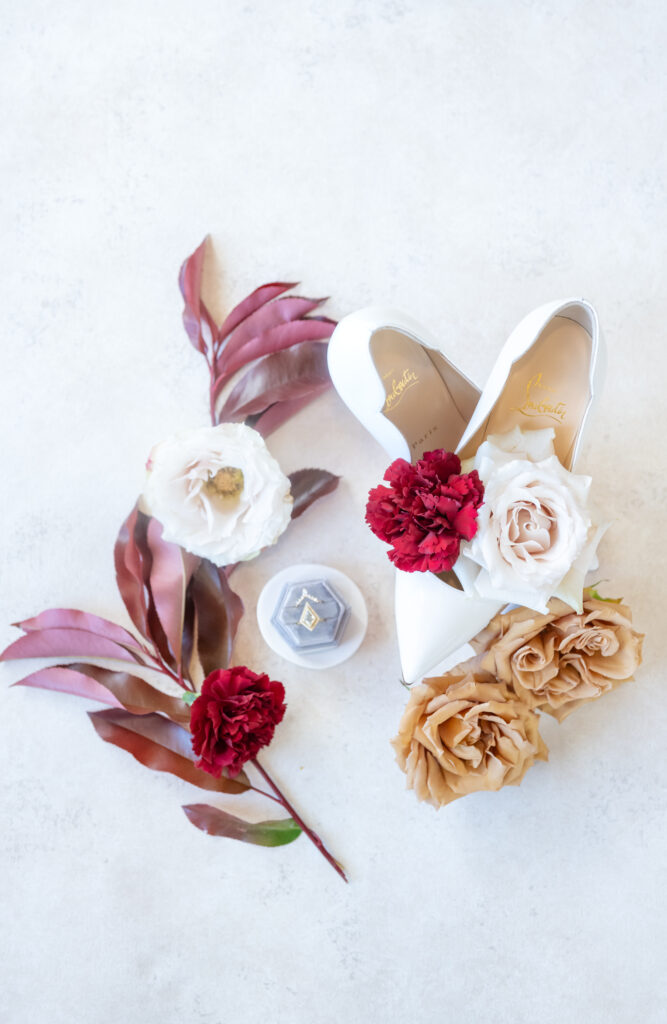Flat lay of bridal wedding details featuring white heels, red and neutral florals, and a wedding ring for a winter wedding at The Broadmoor in Colorado Springs