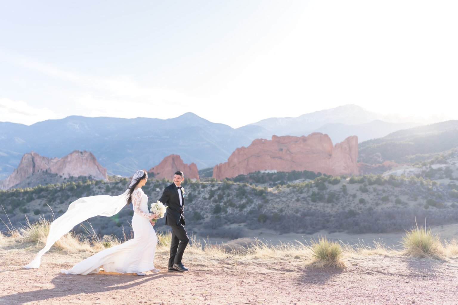 wide shot of bride and groom walking while smiling at each other, red mountains in the background, during their outdoor wedding in colorado springs
