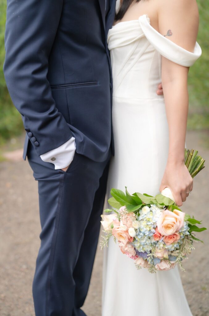 Close-up portrait from a wedding in Colorado Springs showing a bride and groom standing side by side outdoors. The groom wears a tailored navy blue suit with a white dress shirt, his hand tucked casually into his pocket. The bride wears a modern off-the-shoulder ivory wedding gown with a softly draped neckline. She holds a round bouquet filled with blush roses, pale peach blooms, soft blue hydrangeas, and greenery cascading naturally toward the ground. A small heart tattoo is visible on her upper arm. The background is softly blurred greenery, creating an intimate, romantic moment captured by a wedding photographer in Colorado Springs