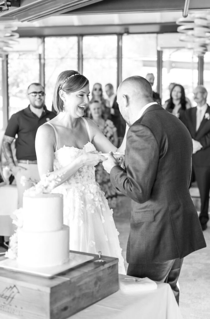 Bride smiling joyfully as she feeds her groom a bite of cake during their reception at a wedding in Colorado Springs. The black and white photograph captures the emotion of the cake cutting moment, with guests watching in the background. A simple two-tier wedding cake sits on a wooden stand, representing elegant wedding cakes in Colorado Springs, while natural window light fills the reception space.