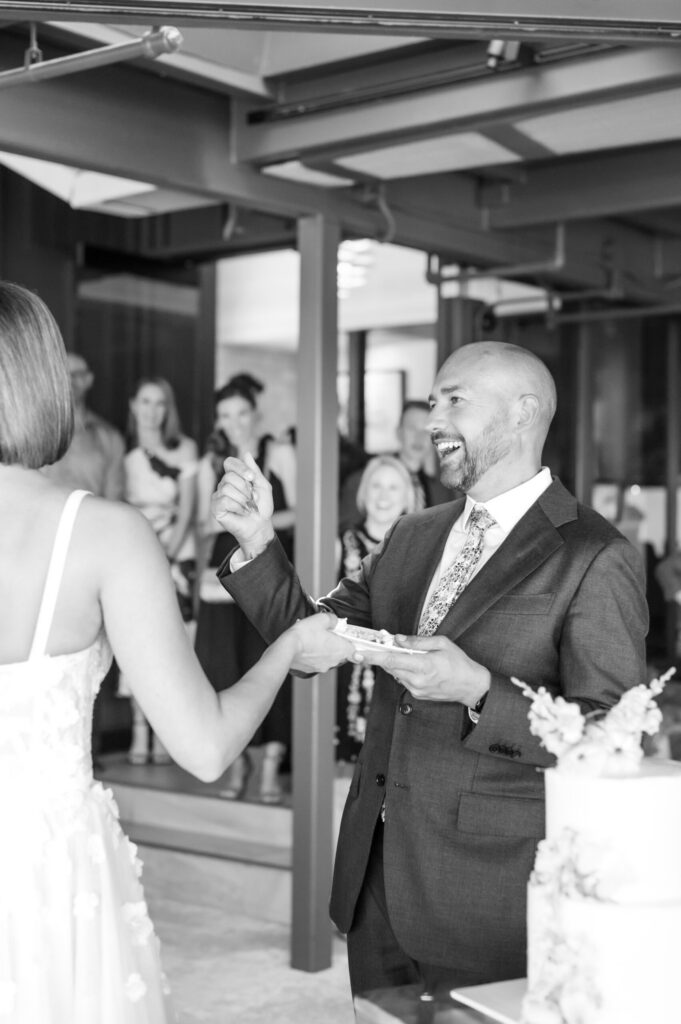 Groom laughing as he holds a small plate of cake during the cake cutting at a wedding in Colorado Springs, surrounded by friends and family inside a modern reception venue. The black and white image highlights the groom’s joyful expression and the celebration atmosphere, showcasing one of the sweetest traditions at weddings in Colorado Springs CO and the artistry behind wedding cakes in Colorado Springs.