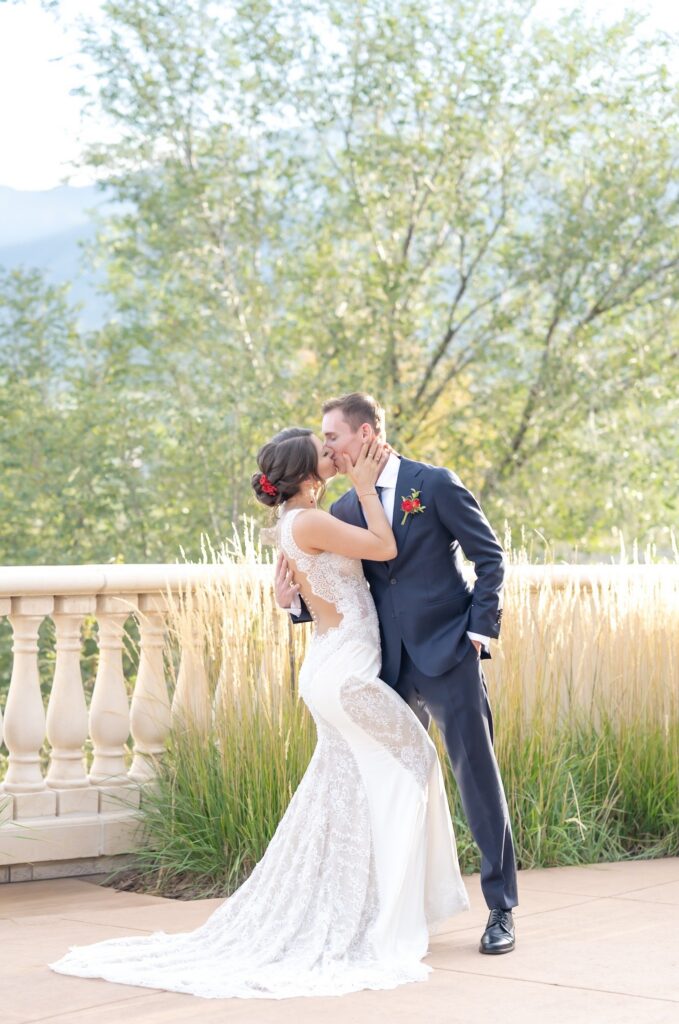 Bride and groom sharing a kiss on a terrace framed by tall ornamental grasses during a romantic sunset wedding in Colorado Springs. The bride’s fitted lace gown features an open back and delicate detailing, while the groom wears a navy suit with a red boutonniere. Soft golden light illuminates the couple, demonstrating why Colorado is known for stunning outdoor wedding locations in Colorado Springs.