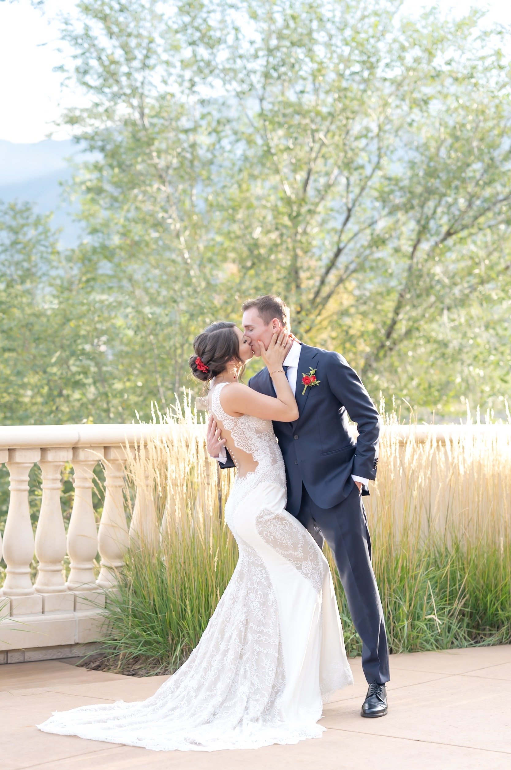 Bride and groom sharing a kiss on a terrace framed by tall ornamental grasses during a romantic sunset wedding in Colorado Springs. The bride’s fitted lace gown features an open back and delicate detailing, while the groom wears a navy suit with a red boutonniere. Soft golden light illuminates the couple, demonstrating why Colorado is known for stunning outdoor wedding locations in Colorado Springs.