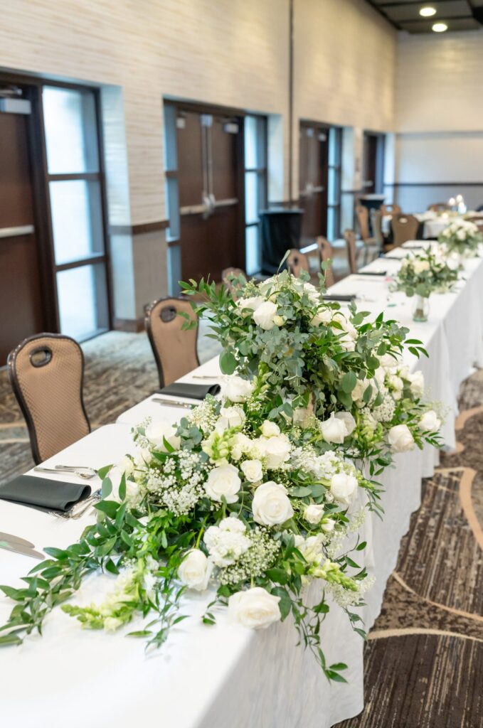 Elegant reception head table adorned with lush white and green floral arrangements, ivory roses, and greenery inside a Colorado Springs wedding venue.