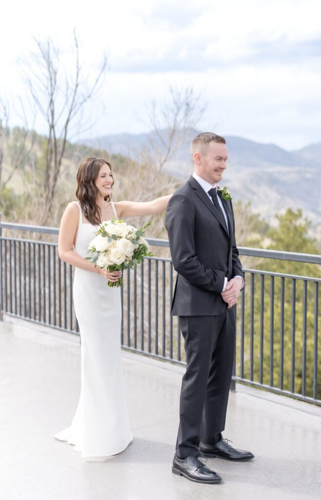 Bride in a fitted white gown holding a bouquet of ivory roses as she taps her groom on the shoulder during their first look on a scenic Colorado Springs mountain overlook with soft blue skies and rolling peaks in the background.