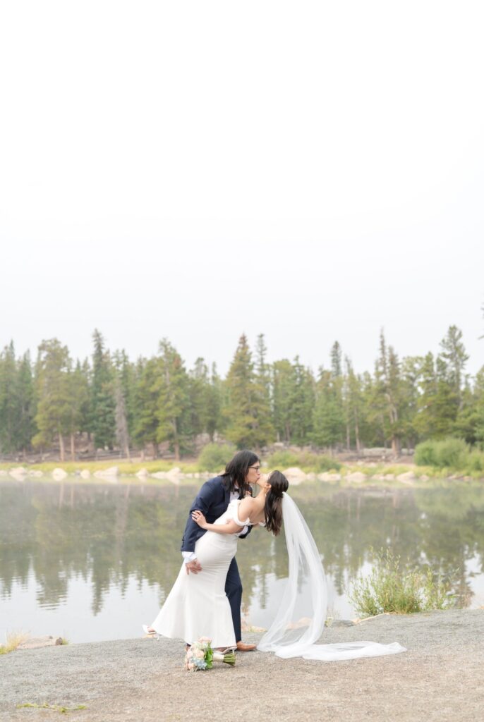 Romantic lakeside dip during a wedding in Colorado Springs, with the groom gently leaning the bride backward for a kiss along the shoreline of a calm mountain lake. Evergreen trees reflect in the still water behind them, and the sky appears softly overcast. The bride’s long veil flows dramatically behind her, trailing across the gravelly lakeside ground. Her bouquet rests near their feet, featuring pastel florals in blush, peach, and blue. This scenic outdoor setting highlights why mountain lakes are among the most breathtaking wedding locations in Colorado Springs and popular places to have weddings in Colorado Springs