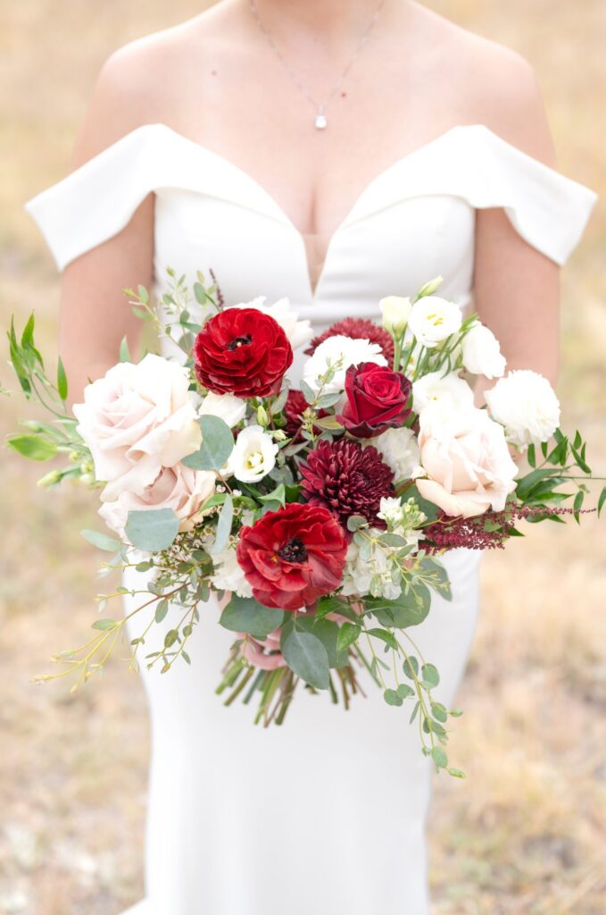 Bride holding a vibrant bouquet of red ranunculus, burgundy mums, blush roses, and greenery against her modern off-the-shoulder wedding gown at a Colorado Springs outdoor wedding.