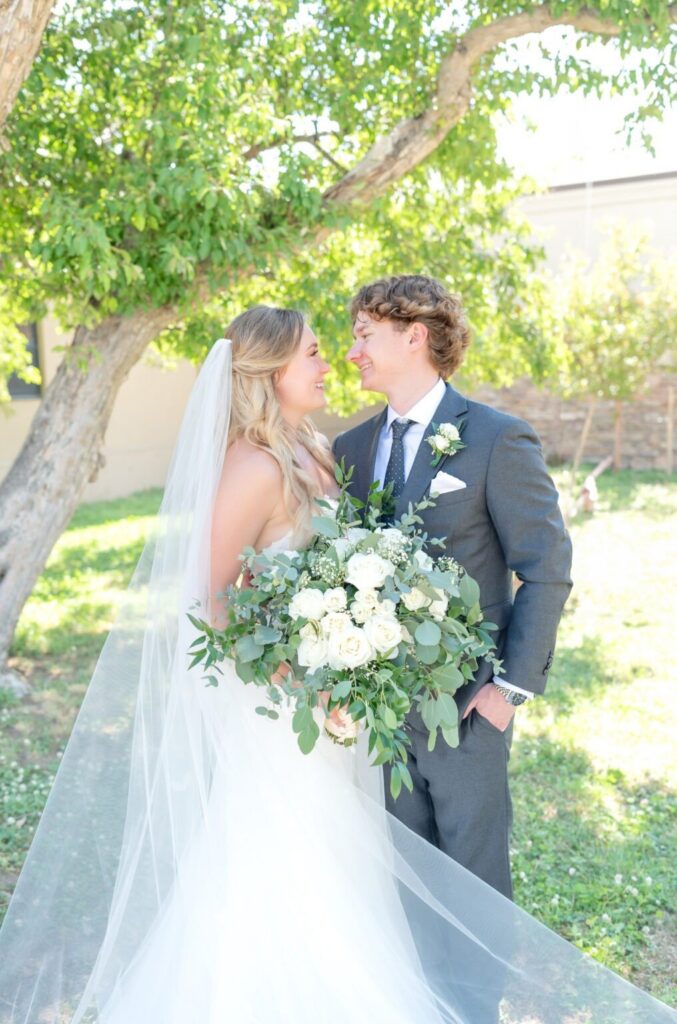 Bride and groom smiling at each other beneath a leafy green tree, the bride holding a lush bouquet of ivory roses and eucalyptus while sunlight filters through the branches.