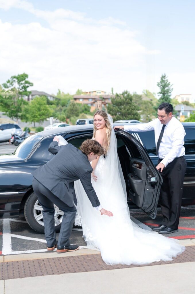 Bride stepping out of a black limousine with her groom adjusting her flowing veil, assisted by a driver, outside a Colorado Springs wedding venue.