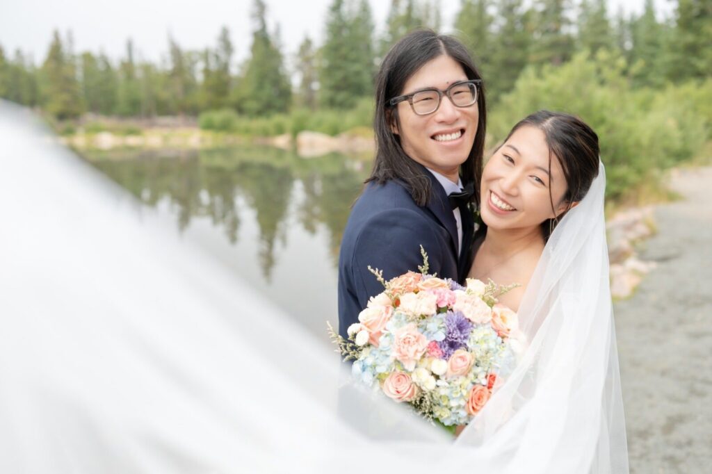 Joyful close-up from a wedding in Colorado Springs featuring a bride and groom embracing near a forested lake. The groom, wearing glasses and a navy suit with a black bow tie, smiles warmly. The bride beams directly at the camera, her veil softly framing her face. She holds a lush pastel bouquet of hydrangeas, roses, and seasonal flowers in soft pink, peach, lavender, and powder blue tones. The calm water and evergreen trees behind them create a peaceful Colorado backdrop, beautifully documented by a Whitney Marshall Photo in Colorado Springs
