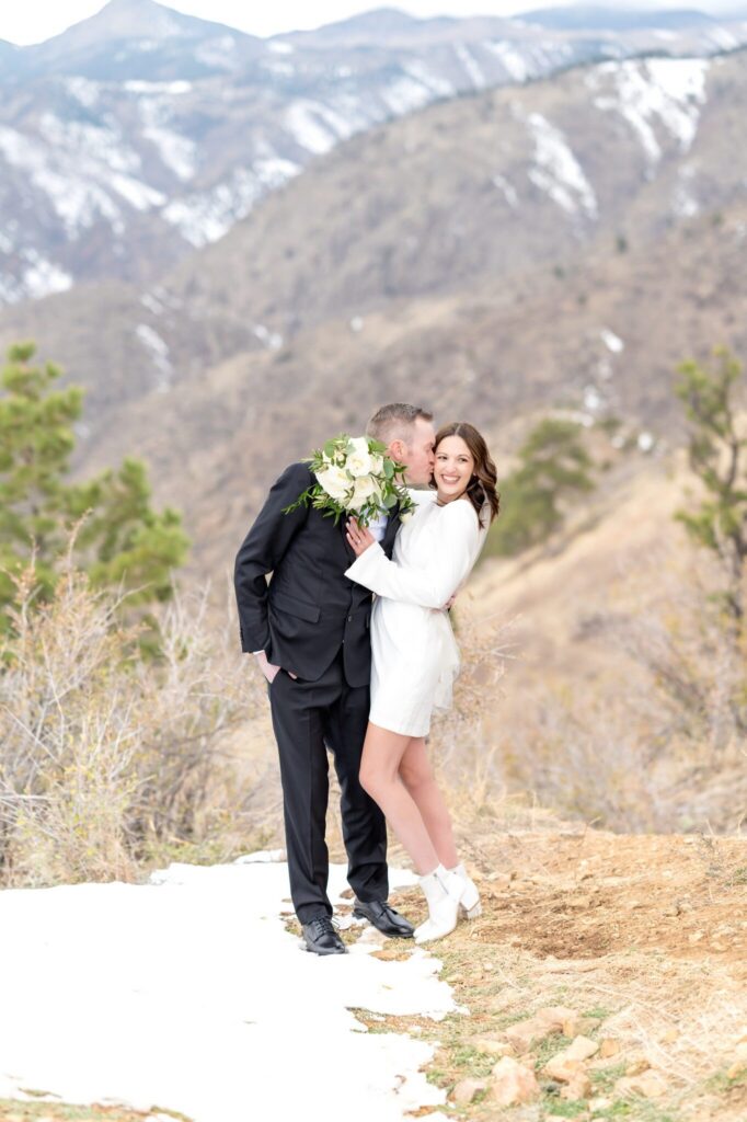Bride in a long-sleeve white mini dress and ankle boots laughing as her groom kisses her cheek on a snowy Colorado mountainside.