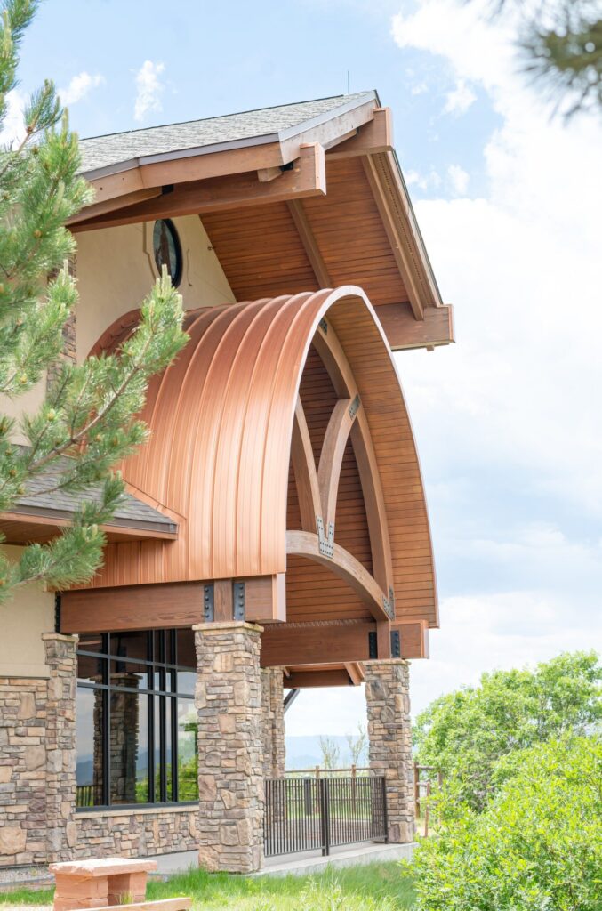 Exterior of a Colorado Springs wedding venue featuring stone columns, large windows, and a curved copper architectural awning framed by green trees.