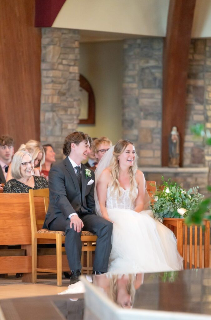 Bride and groom laughing together during their wedding ceremony inside a chapel with stone walls and wooden pews, surrounded by guests.