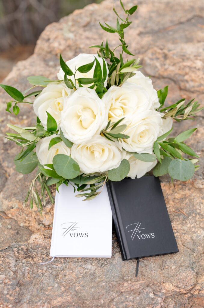 Ivory rose wedding bouquet with lush greenery resting on natural stone beside black and white personalized vow books during a Colorado Springs mountain elopement.