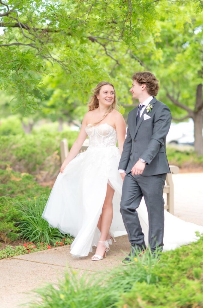 Bride and groom walking hand in hand along a garden path, the bride’s flowing gown and veil moving softly as they smile at each other beneath vibrant green trees.