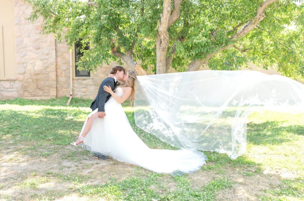 Romantic portrait of the bride and groom sharing a kiss under a tree, the bride’s long veil flowing dramatically behind her across the grass.