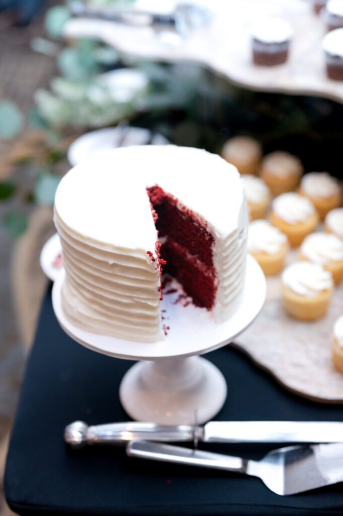 Close-up of a single-tier red velvet wedding cake with smooth white buttercream frosting and a slice removed, displayed on a white cake stand with cupcakes blurred in the background.
