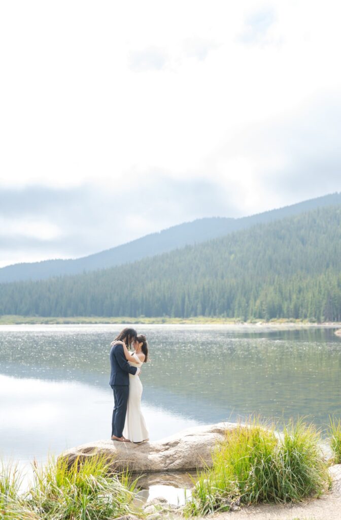 Wide scenic view of a wedding in Colorado Springs with a bride and groom standing together on a large natural rock beside a glassy alpine lake. Pine-covered mountains rise in the distance beneath a bright sky. The couple stands close, foreheads nearly touching, wrapped in a quiet embrace. The bride’s fitted gown falls elegantly to the rock beneath her, and her long veil drapes gently in the breeze. This expansive natural landscape showcases why mountain lakes are among the most sought-after wedding locations in Colorado Springs and unforgettable places to have weddings in Colorado Springs