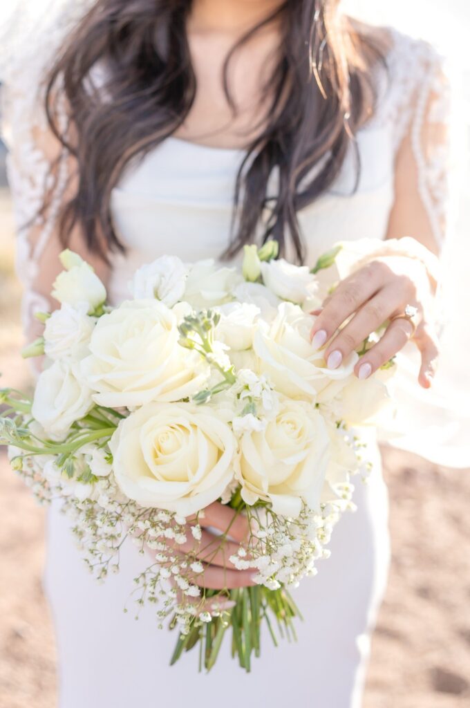 Bride holding a rounded bouquet of ivory roses and delicate white blooms, her long dark hair and lace sleeves softly backlit during a Colorado Springs elopement.
