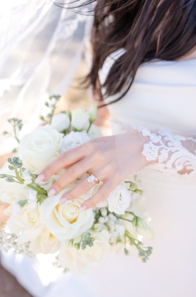 Close-up of bride’s hand with oval diamond engagement ring resting on a bouquet of ivory roses and white florals, lace sleeve softly illuminated by golden sunlight.