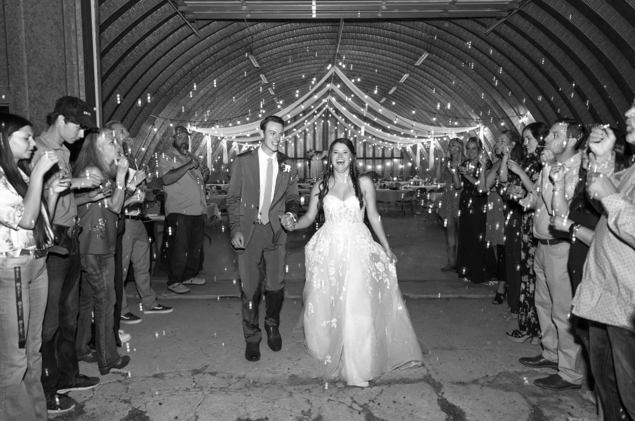 Black and white photo of bride and groom walking hand in hand through a sparkler exit inside a rustic barn reception space, surrounded by cheering guests under string lights on their Colorado wedding day.