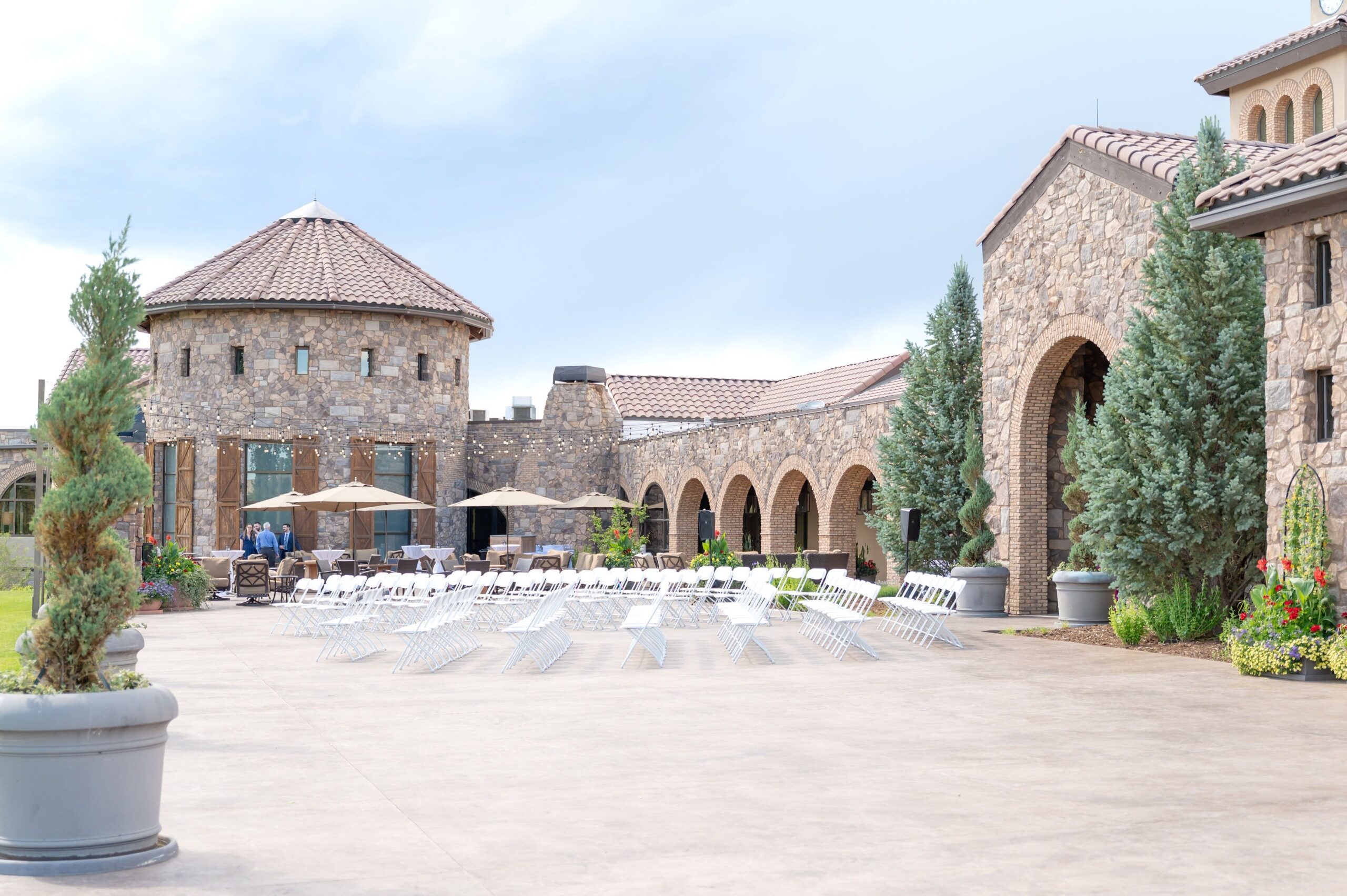 Outdoor wedding ceremony setup at a Colorado Springs stone venue featuring arched walkways, a round tower, and rows of white folding chairs arranged on a spacious courtyard patio.