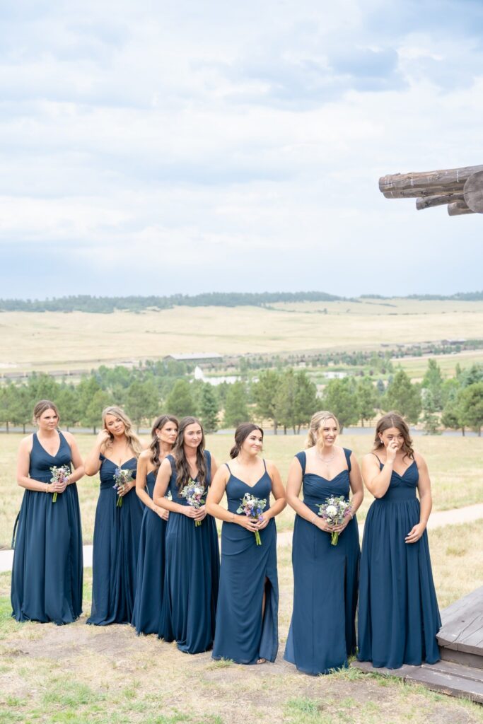 Bridesmaids in matching navy blue floor-length dresses holding small bouquets during an outdoor Colorado wedding ceremony, with open fields and mountain views behind them.