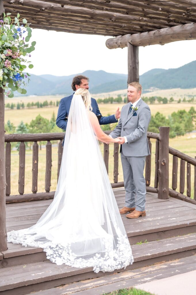 Bride and groom holding hands during an outdoor wedding ceremony under a wooden pergola at Spruce Mountain Ranch, overlooking the Colorado countryside and distant mountains.