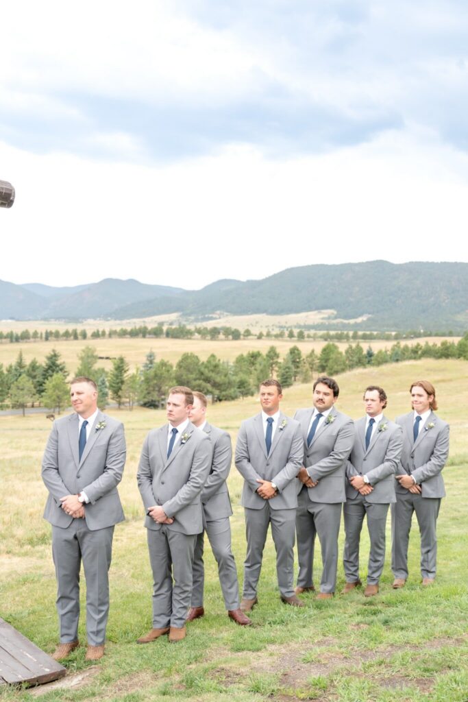 Groomsmen in light gray suits with navy ties standing in a row during an outdoor Colorado wedding ceremony, with rolling hills, pine trees, and mountain views in the background.