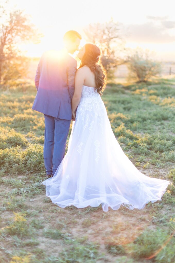 Bride and groom holding hands during golden hour portraits in a Colorado field, with warm sunset light glowing around them and the bride’s flowing gown trailing behind.