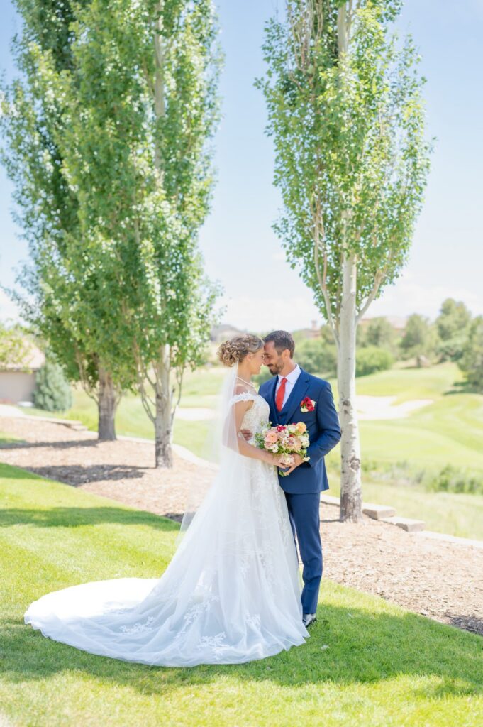 Bride and groom standing closely together between two tall, slender trees on a manicured golf course during a wedding in Colorado Springs. The bride wears a lace-detailed gown with a flowing train spread across the vibrant green grass. The groom wears a navy blue suit accented with a bold red tie and matching boutonniere. Soft hills and fairways stretch into the distance under bright sunlight. The couple gazes at one another while holding a bouquet of warm-toned flowers. This outdoor golf course venue is one of the elegant places to have weddings in Colorado Springs