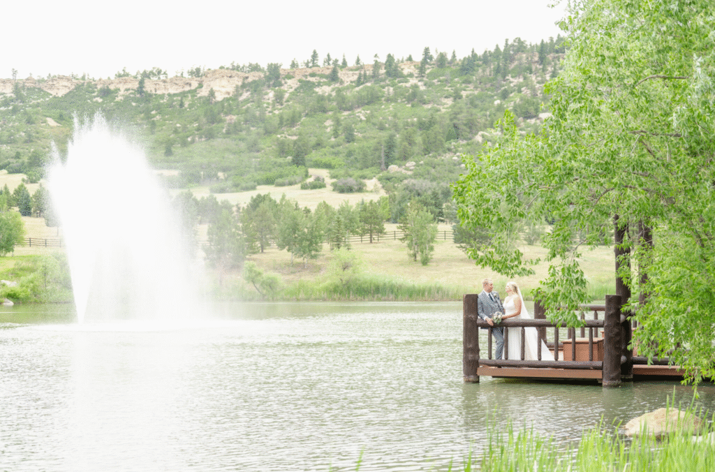 Bride and groom standing together on a rustic wooden dock extending over a peaceful lake during a wedding in Colorado Springs. A large fountain sprays water into the air on the left side of the frame, creating a dramatic mist against the calm surface. Rolling green hills and scattered trees stretch into the background. The bride holds a bouquet while facing the groom, both framed by leafy branches overhead. This elegant waterfront setting represents one of the many picturesque wedding locations in Colorado Springs and highlights the variety of weddings in Colorado Springs CO