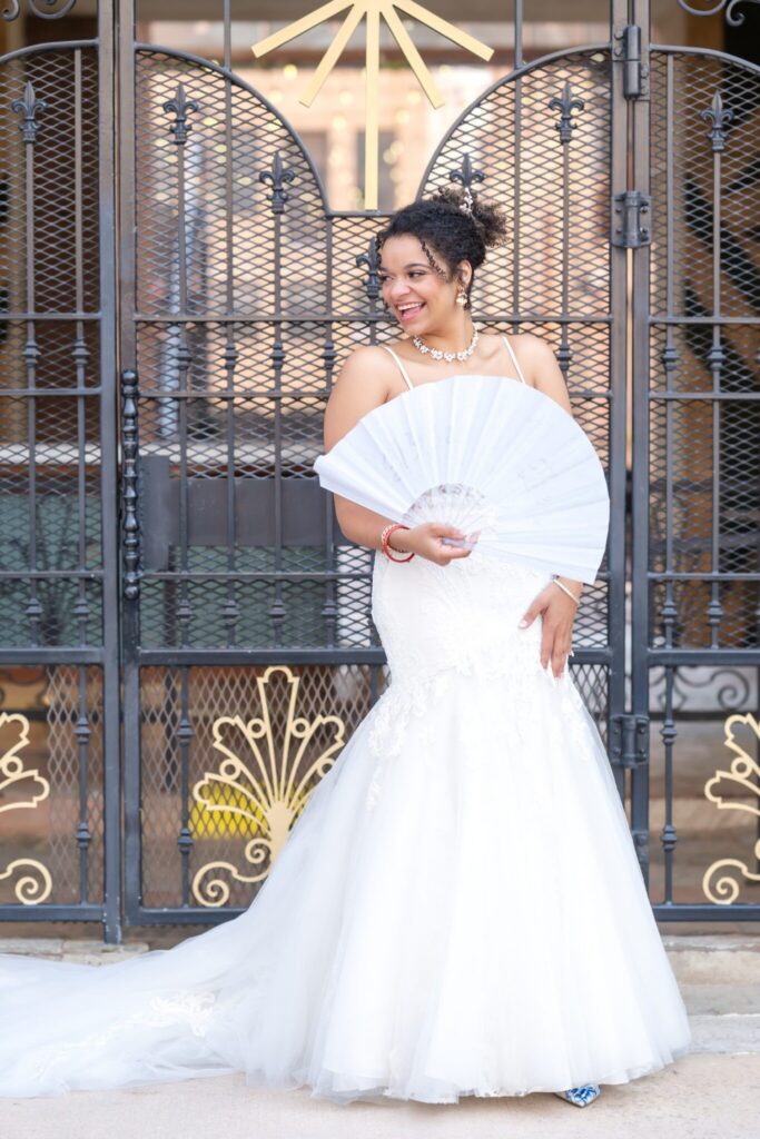 Bride smiling and holding a white folding fan in front of ornate wrought iron gates during an elegant wedding in Colorado Springs. She wears a fitted lace wedding gown with a soft tulle skirt and delicate floral detailing, a sparkling choker necklace, and her hair styled in a curly updo. The decorative iron doors behind her feature gold accents and intricate patterns, creating a romantic architectural backdrop often found at wedding locations in Colorado Springs. This portrait captures modern bridal style and refined venue details from weddings in Colorado Springs CO, beautifully documented by a wedding photographer in Colorado Springs