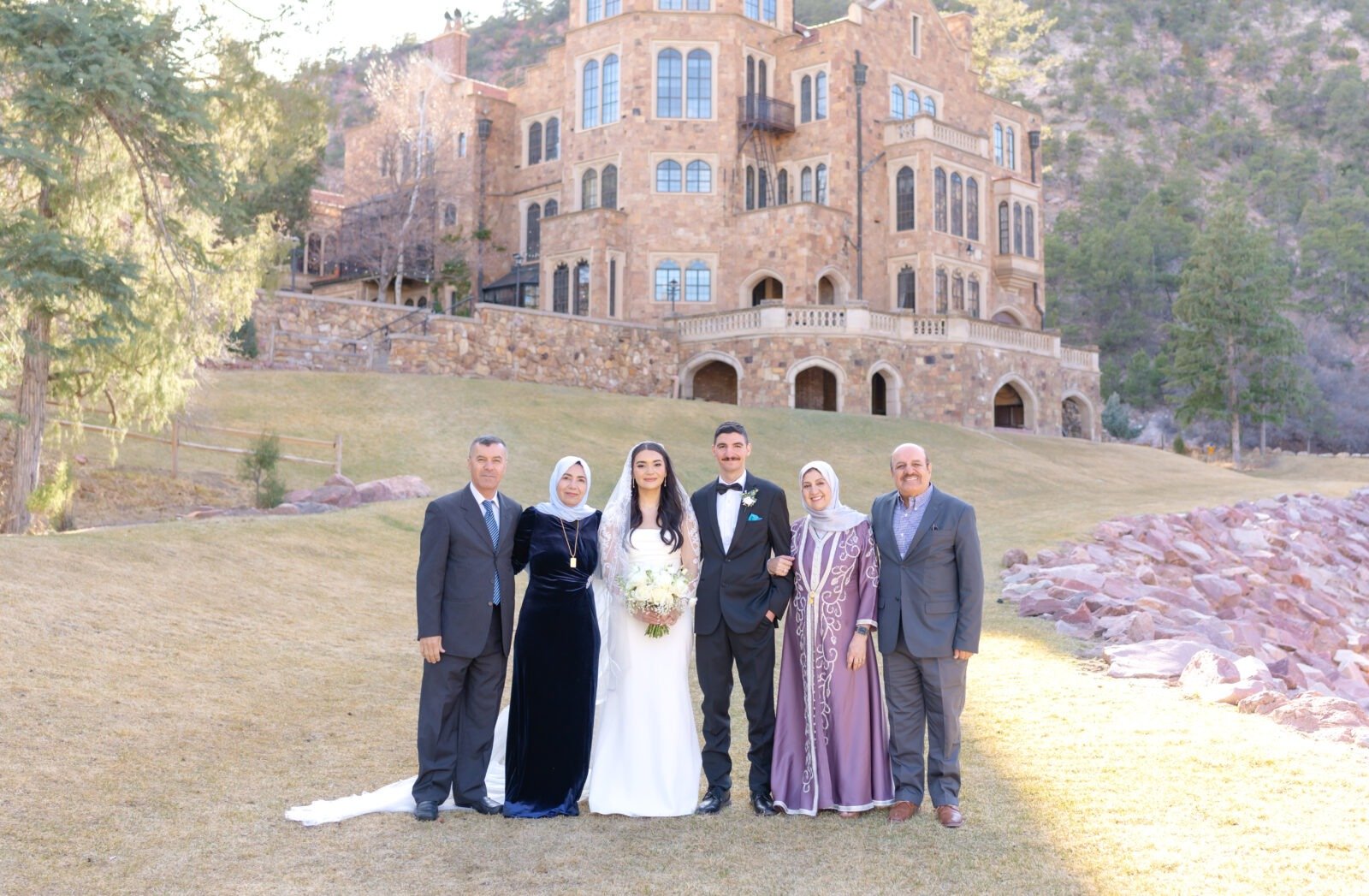 Bride and groom standing with family members on a grassy lawn in front of a grand stone estate during a wedding in Colorado Springs. The castle-style venue features arched windows, balconies, and textured stone walls set against foothills and evergreen trees, highlighting one of the most scenic places to have weddings in Colorado Springs. The bride wears a classic white gown with a long veil and holds a bouquet of white flowers, while family members stand beside them in formal attire, including elegant dresses and tailored suits. This timeless family portrait reflects multicultural celebration and showcases one of the premier wedding locations in Colorado Springs